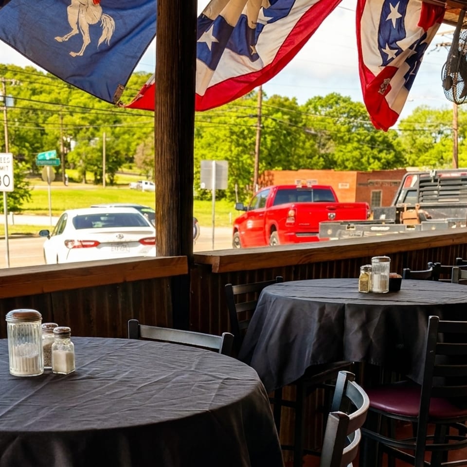 Outdoor patio dining area with black-covered tables and chairs, overlooking a street with cars and trees.