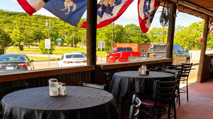 Outdoor patio dining area with black-covered tables and chairs, overlooking a street with cars and trees.