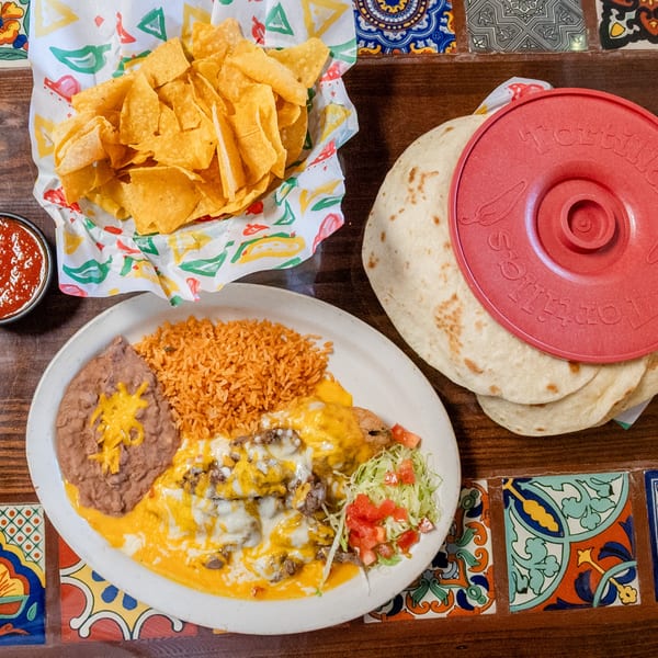 Mexican dinner with chips, salsa, cheesy meat entree, rice, beans, and warm tortillas on a tiled table.