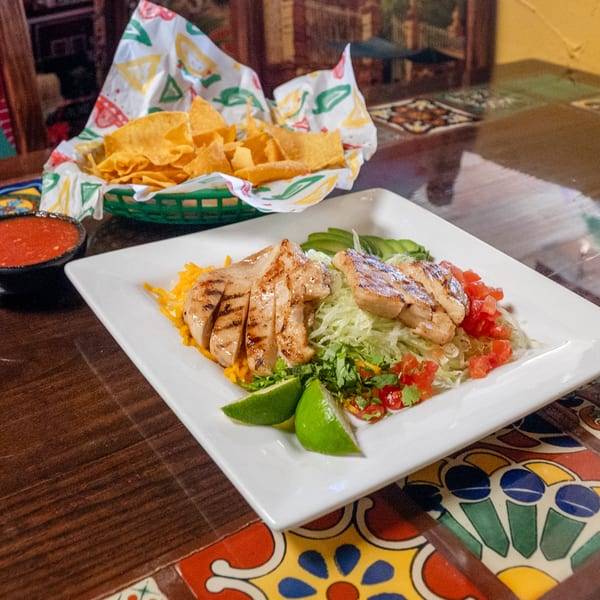 Grilled chicken salad with avocado, lime, and salsa, served with tortilla chips on a tiled table.