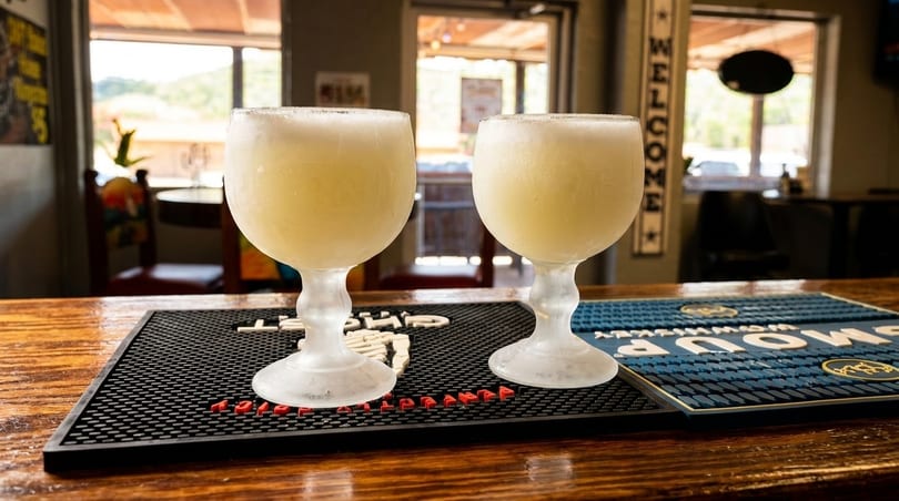 Two frosty margarita glasses filled with a pale yellow drink sit on a bar mat.