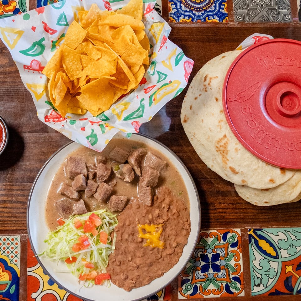 Mexican meal with beef stew, refried beans, chips, salsa, and warm tortillas on a colorful tiled table.