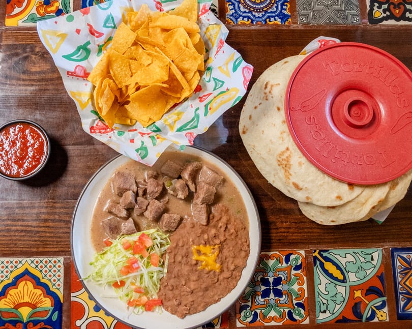 Mexican meal with beef stew, refried beans, chips, salsa, and warm tortillas on a colorful tiled table.