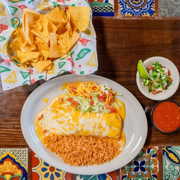Cheese-covered burrito with rice, tortilla chips, salsa, and pico de gallo on a tiled wooden table.