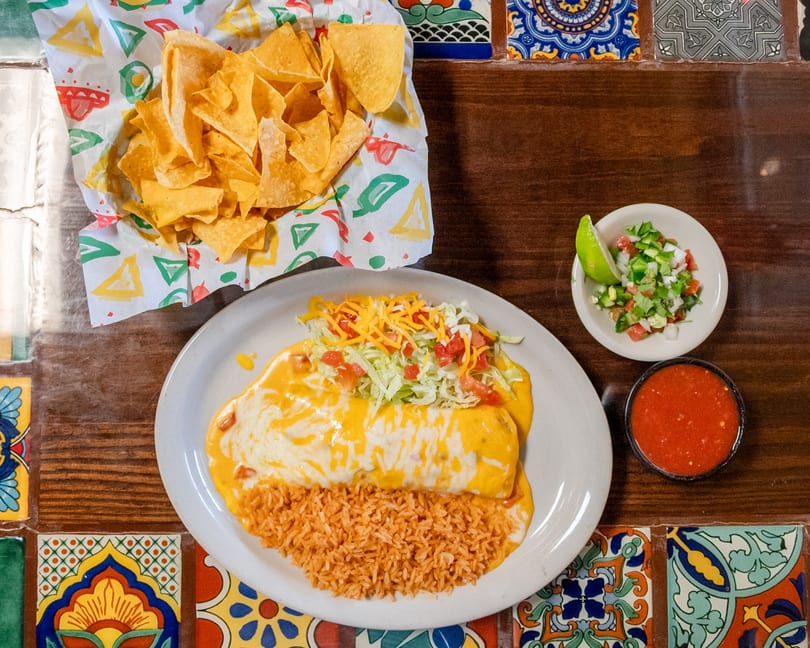 Cheese-covered burrito with rice, tortilla chips, salsa, and pico de gallo on a tiled wooden table.