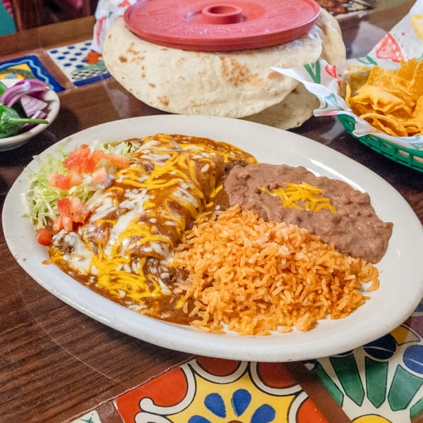 Mexican dinner plate with cheesy enchiladas, rice, beans, tortillas, chips, and salsa on a vibrant tiled table.