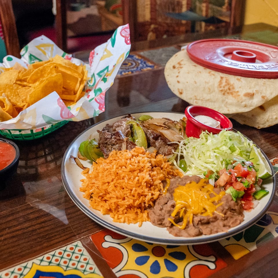 Fajita plate featuring steak, peppers, onions, rice, refried beans, lettuce, pico de gallo, chips, salsa, and tortillas.