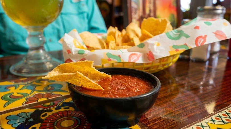 Tortilla chips in a basket and a bowl of salsa with chips, on a colorful tiled table.