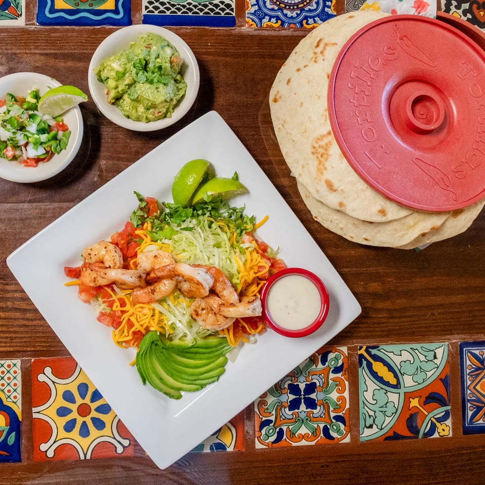 Grilled shrimp fajita plate with guacamole, pico de gallo, and tortillas on a tiled wooden table.