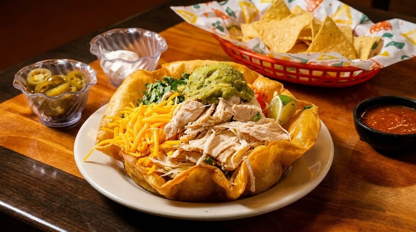 Chicken taco salad in a fried tortilla bowl with guacamole, cheese, and cilantro, served with chips and salsa.