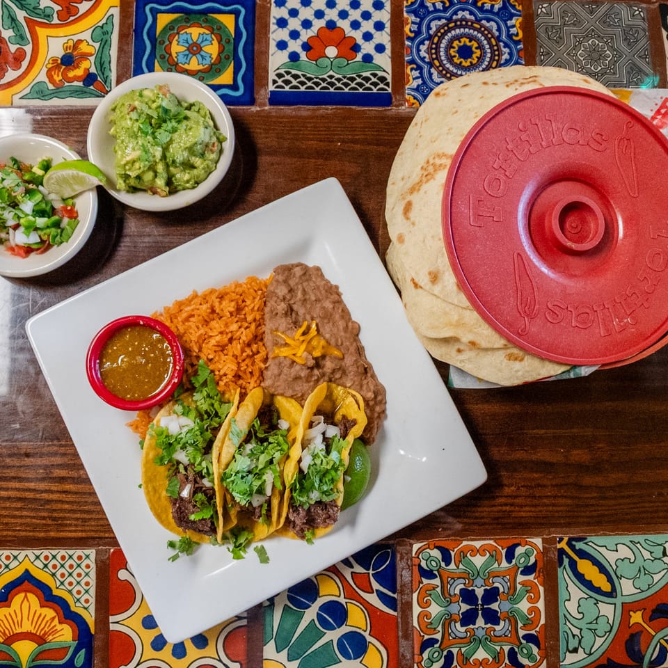 Plate of tacos with rice, beans, salsa, guacamole, pico de gallo, and tortillas on a tiled table.