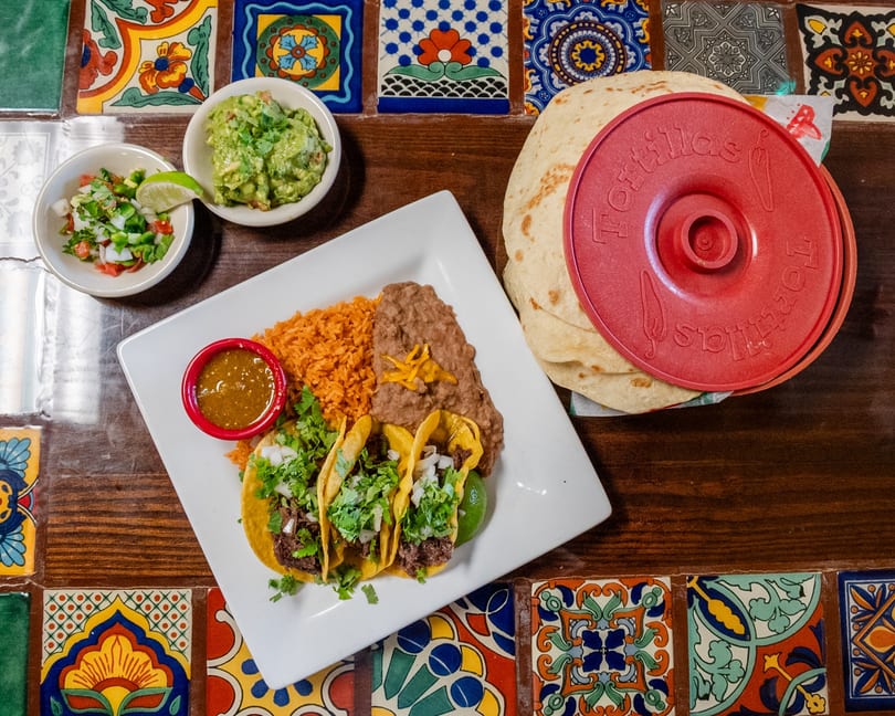 Plate of tacos with rice, beans, salsa, guacamole, pico de gallo, and tortillas on a tiled table.