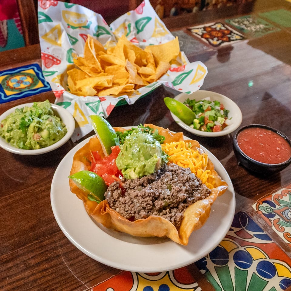 Colorful taco salad with ground beef, guacamole, and cheese, alongside chips, salsa, and pico de gallo on a tiled table.