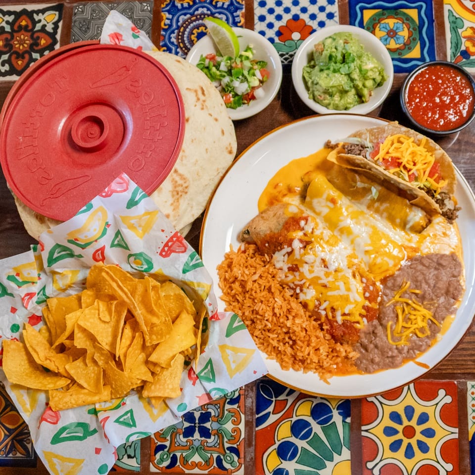 Mexican combo plate with enchiladas, rice, beans, taco, chips, salsa, guacamole, and tortillas on a colorful tiled table.