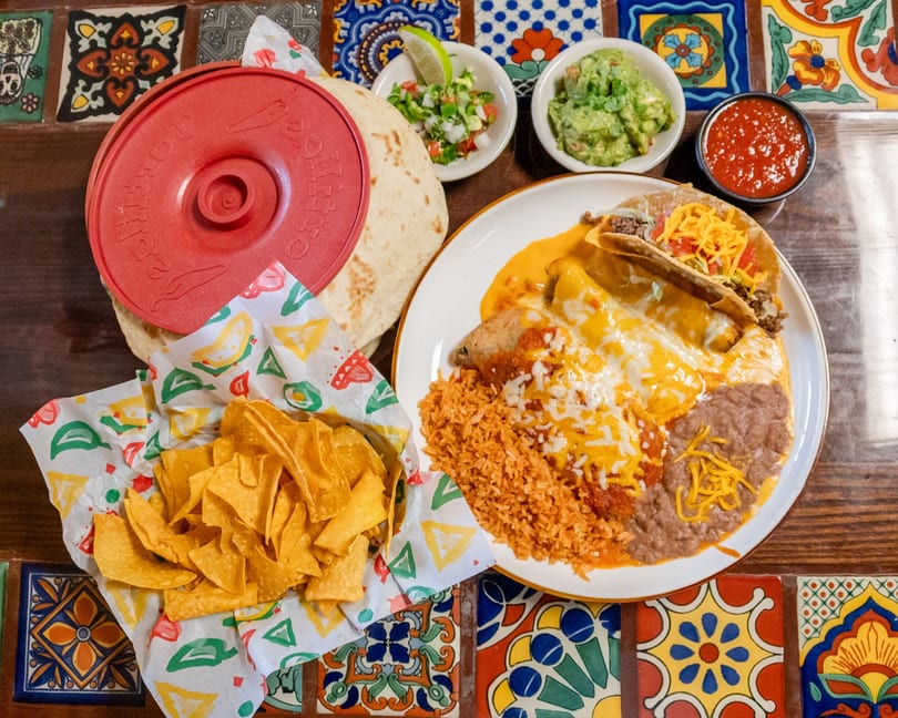 Mexican combo plate with enchiladas, rice, beans, taco, chips, salsa, guacamole, and tortillas on a colorful tiled table.