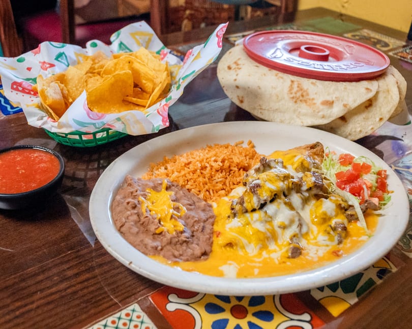 Platter of cheesy Mexican entree with rice, refried beans, chips, salsa, and warm tortillas.