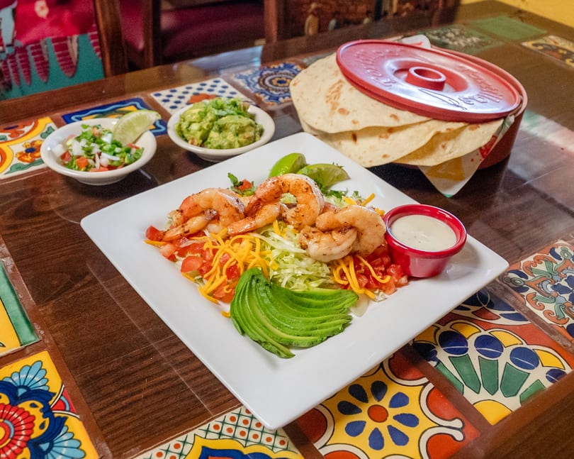 Shrimp fajita plate with guacamole, pico de gallo, and tortillas on a colorful tiled table.
