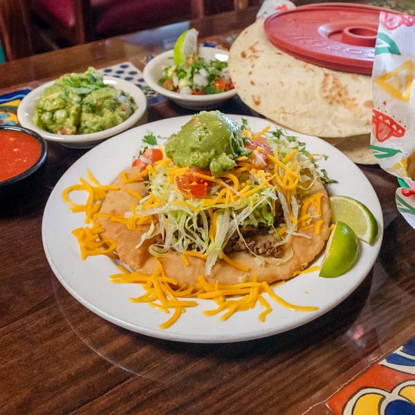 A large, loaded taco with meat, cheese, lettuce, tomatoes, and guacamole, served with salsa, chips, and tortillas.