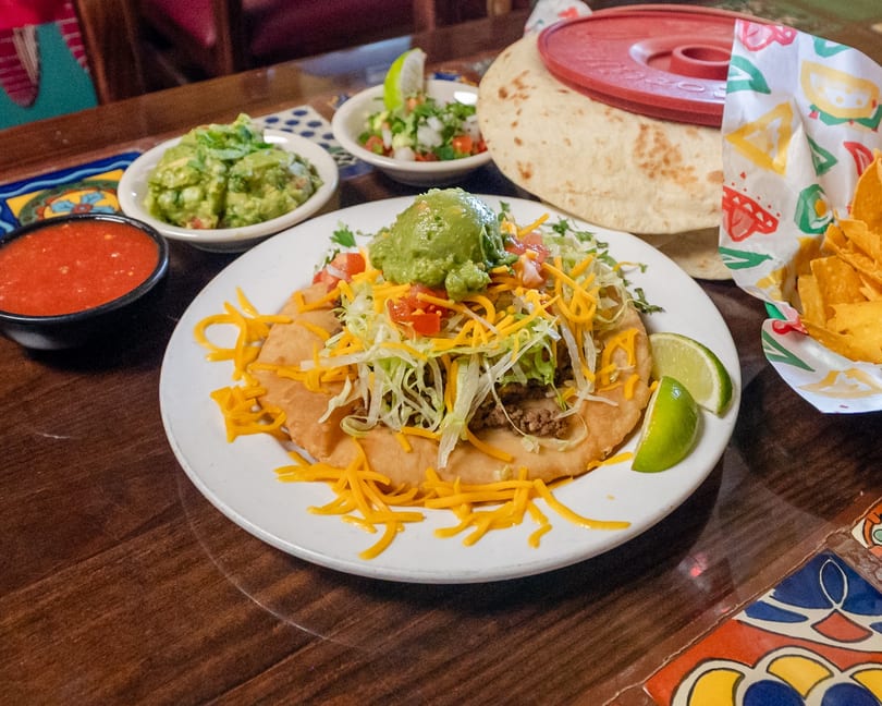 A large, loaded taco with meat, cheese, lettuce, tomatoes, and guacamole, served with salsa, chips, and tortillas.