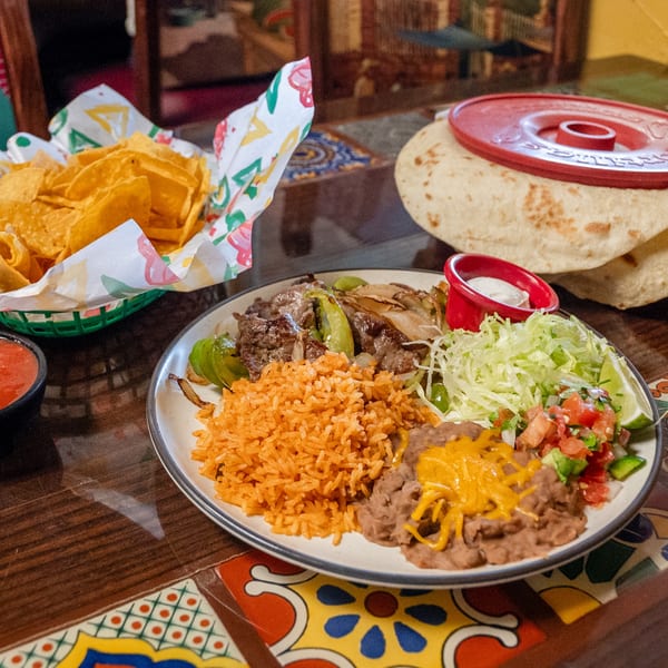 Plate of steak fajitas with rice, beans, lettuce, pico, tortillas, chips, and salsa on a tiled table.