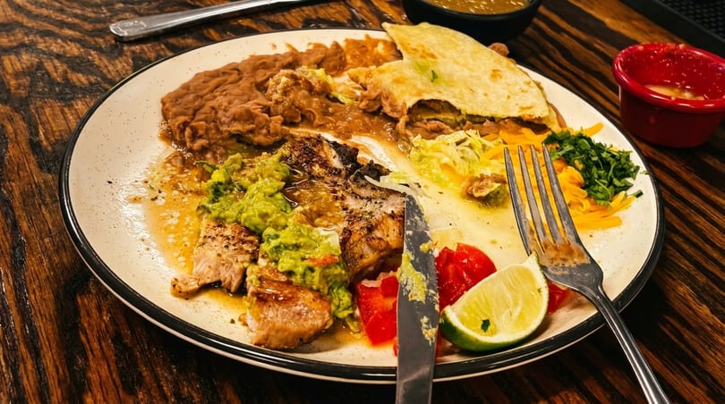 A partially eaten plate of Mexican food with grilled meat, guacamole, refried beans, and a quesadilla.