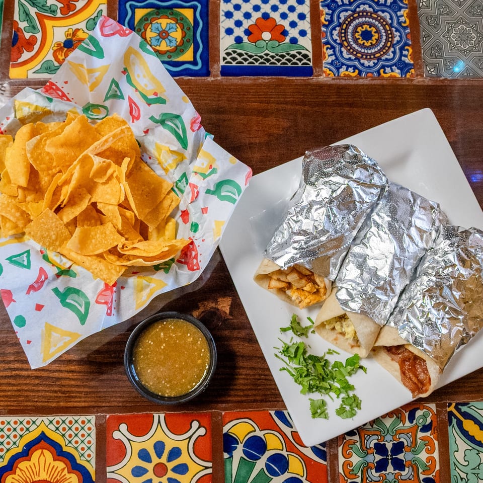 Three burritos, a basket of tortilla chips, and green salsa on a colorful tiled table.