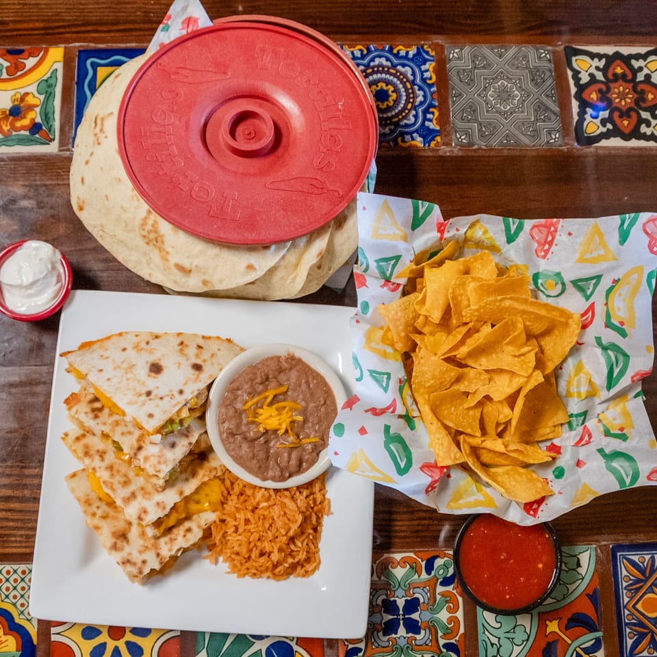 Quesadillas with rice, beans, chips, salsa, sour cream, and tortillas on a colorful tiled table.